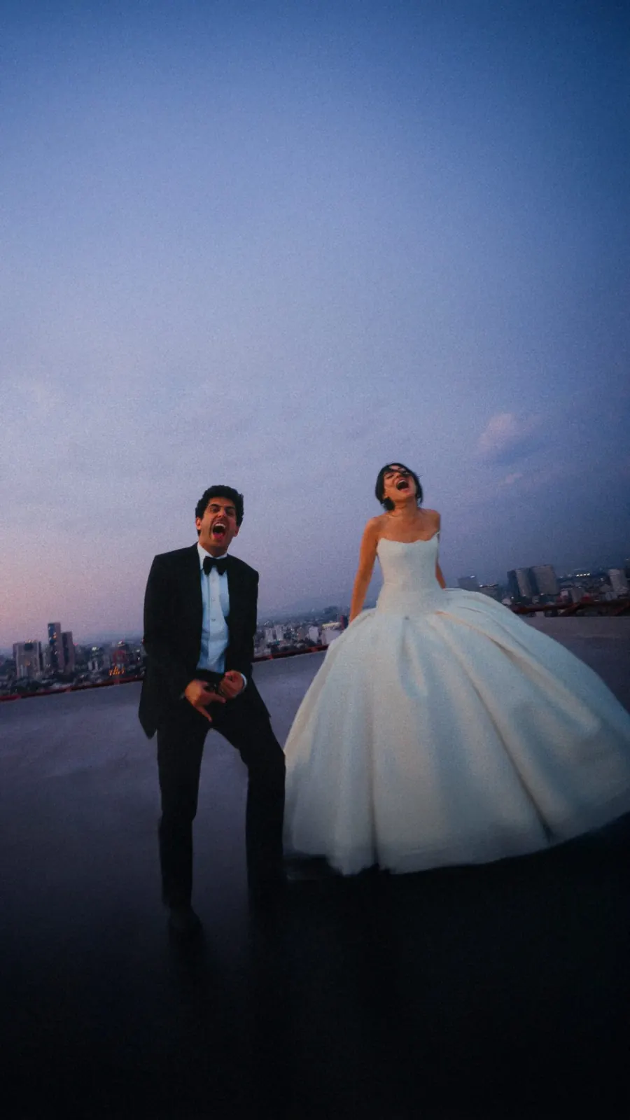 Bride and groom laughing on a rooftop at dusk with the city skyline behind them.